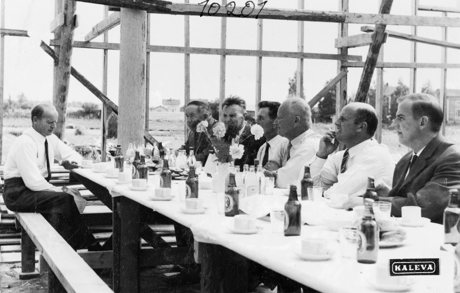 Miehiä pitkän pöydän ääressä harjannostajaisissa rakennustyömaalla vuonna 1961